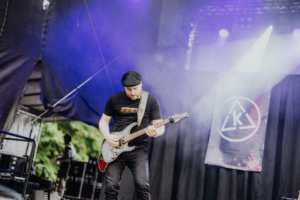 Kontrollverlust Ein Musiker spielt auf der Bühne des Elbenwald Festivals Gitarre. Im Hintergrund sind Lichteffekte und ein Banner sichtbar, das das Festival-Logo zeigt. Die Atmosphäre ist energiegeladen und zieht ein nerdiges Publikum an.