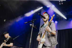 Kontrollverlust Ein Sänger mit buntem Haar und Gitarre steht auf der Bühne des Elbenwald Festivals. Er trägt ein gestreiftes T-Shirt und eine Jeansweste. Im Hintergrund sind Lichter und eine schwarze Bühne zu sehen, die die lebendige Atmosphäre des Festivals unterstreichen.