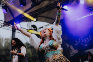 Kupfergold Eine fröhliche Sängerin mit buntem Kostüm und Blumen im Haar begeistert das Publikum auf der Bühne des Elbenwald Festivals, umgeben von bunten Lichtern und einer lebhaften Atmosphäre. Im Hintergrund ist ein Musiker mit Gitarre zu sehen.