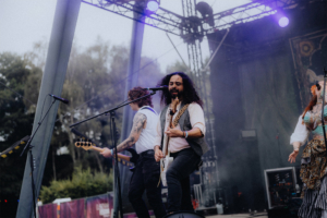 Kupfergold Musiker auf der Bühne des Elbenwald Festivals, umgeben von bunten Lichtern und Nebel. Die Band spielt energetisch, während das Publikum begeistert zuschaut. Die Atmosphäre vereint Musik und Fantasie in einer einzigartigen Festivalerfahrung.