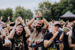 Kupfergold Menschenmenge beim Elbenwald Festival, die begeistert applaudiert. Verschiedene Kostüme und Outfits, die die nerdige Kultur widerspiegeln. Eine lebendige Atmosphäre mit fröhlichen Festivalbesuchern in einer grünen Umgebung.