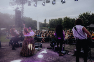 Kupfergold Blick von der Bühne beim Elbenwald Festival, mit einer Sängerin in traditioneller Kleidung und Bandmitgliedern, die vor einer begeisterten Menge auftreten. Im Hintergrund ist eine grüne Landschaft und ein Wasserfall zu sehen, während die Lichter auf die Bühne gerichtet sind.