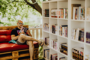 Carlsen Manga Lounge Ein Mann sitzt entspannt auf einer roten Couch aus Paletten und liest ein Manga-Heft. Im Hintergrund steht ein Bücherregal mit einer Vielzahl von Manga-Bänden. Die Szene vermittelt eine gemütliche Atmosphäre auf dem Elbenwald Festival.