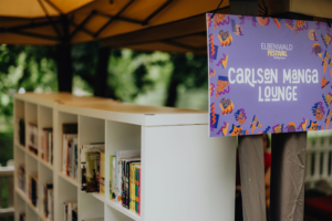 Carlsen Manga Lounge Ein farbenfrohes Schild der Carlsen Manga Lounge auf dem Elbenwald Festival, das eine moderne Buchauslage mit Manga und Comics zeigt, umgeben von einer entspannten Festivalatmosphäre und Bäumen im Hintergrund.