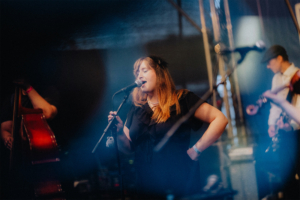 Mardi Sängerin auf der Bühne beim Elbenwald Festival, umgeben von Bandmitgliedern. Die Atmosphäre ist lebhaft und festlich, während das Publikum die musikalische Darbietung genießt. Farbige Lichter und Instrumente schaffen eine einladende Festivalstimmung.