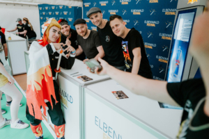 Kontrollverlust Cosplayer und Besucher am Elbenwald Festival, posierend an einem Stand mit Merchandise. Im Hintergrund sind Festivalbanner sichtbar. Die Atmosphäre ist lebhaft und freundlich, ideal für Fans von Musik und Popkultur.