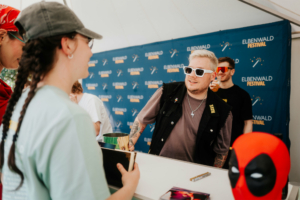 Meet and Greet Nerds at Raves Besucher am Stand des Elbenwald Festivals, lächelnd im Gespräch mit einem Mitarbeiter. Im Hintergrund ist das Festival-Logo sichtbar. Der Stand bietet Merchandise und Informationen zu den Veranstaltungen und Aktivitäten des Festivals.