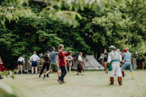 Workshop Bild: Gina WetzlerSchwertkampf Teilnehmer des Elbenwald Festivals in fantasievollen Kostümen üben mit Stöcken im Freien. Die Veranstaltung kombiniert Musik, Rollenspiel und Gemeinschaftsaktivitäten in einer lebhaften, grünen Umgebung.