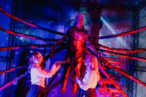 Stranger Things Zwei Cosplayerinnen interagieren mit einer beeindruckenden, mehrarmigen Statue in einer bunten, beleuchteten Umgebung beim Elbenwald Festival. Die Szenerie kombiniert Elemente von Musik und Fantasy und zieht zahlreiche Nerds und Fans an.