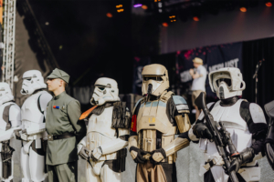 501st Legion Gruppierung von Stormtrooper-Kostümen und einem imperialen Offizier auf der Bühne des Elbenwald Festivals, umgeben von festlicher Atmosphäre, während ein Musiker im Hintergrund auftritt. Ideal für Fans von Science-Fiction und Cosplay.