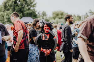 Eine Gruppe von Festivalbesuchern beim Elbenwald Festival, darunter eine Person in auffälligem Teufelskostüm mit roter Gesichtsbemalung und einem großen, dekorativen Stock. Die Atmosphäre ist lebhaft und bunt, mit vielen anderen kostümierten Teilnehmern im Hintergrund.