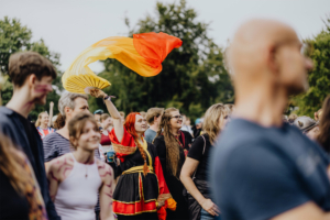 Menschenmenge beim Elbenwald Festival, mit einer festlichen Teilnehmerin in buntem Kostüm, die einen farbenfrohen Fächer schwingt. Die Atmosphäre ist lebhaft und voller Enthusiasmus, während die Besucher die Musik und das Gemeinschaftsgefühl genießen.