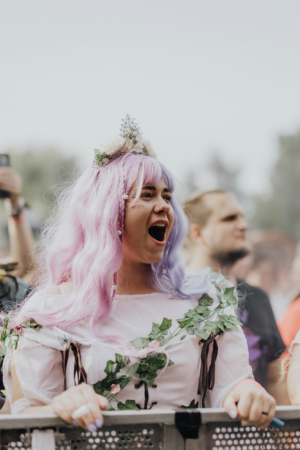 Junge Frau mit lila Haaren und Blumenkranz, die begeistert auf dem Elbenwald Festival steht. Sie trägt ein Kostüm mit Pflanzenmotiven und zeigt ihre Freude während eines Auftritts, umgeben von einer lebhaften Festivalatmosphäre.