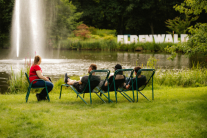 Besucher des Elbenwald Festivals entspannen in Liegestühlen am Ufer eines Teiches, während im Hintergrund das Festival-Logo zu sehen ist. Die Szene zeigt eine ruhige Atmosphäre umgeben von Natur und Wasser.