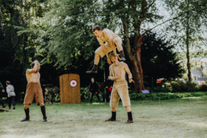 Ompah Kunstreitshow Drei Darsteller in fantasievoller Kleidung führen eine akrobatische Darbietung auf dem Elbenwald Festival auf. Im Hintergrund ist eine Zielscheibe zu sehen, während Zuschauer die spannende Performance beobachten.