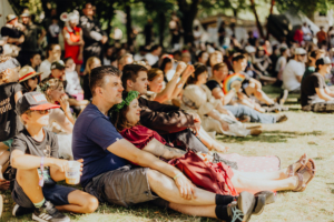 Besucher des Elbenwald Festivals genießen die Atmosphäre auf der Wiese, während sie einer Aufführung auf der Bühne lauschen. Die bunte Mischung aus Cosplay und Musik schafft eine lebendige, gemeinschaftliche Stimmung unter den Festivalteilnehmern.