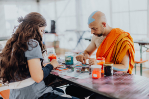Retro-Basteln Zwei Teilnehmer des Elbenwald Festivals arbeiten an Bastelprojekten. Eine Person trägt ein Kostüm mit langen Haaren und Brille, während die andere einen orangefarbenen Umhang und einen blauen Strich auf der Stirn hat. Verschiedene Bastelmaterialien sind auf dem Tisch verteilt.