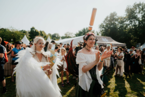 Zwei Frauen in fantasievollen Kostümen applaudieren fröhlich beim Elbenwald Festival. Im Hintergrund sind andere Festivalbesucher und Zelte zu sehen, die die lebendige Atmosphäre des Events unterstreichen. Ein unvergessliches Erlebnis für Fantasy-Fans.