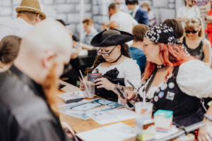 Teilnehmer des Elbenwald Festivals sitzen an einem Tisch, arbeiten an kreativen Projekten und genießen Getränke. Die Atmosphäre ist lebhaft, mit einer Mischung aus Kostümen und Dekorationen, die die nerdige Kultur widerspiegeln.