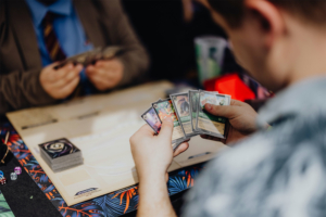 Ein Teilnehmer des Elbenwald Festivals hält mehrere Spielkarten in der Hand, während er an einem Tisch sitzt, umgeben von anderen Spielern. Im Hintergrund sind weitere Karten und Würfel sichtbar, die die spielerische Atmosphäre des Events unterstreichen.