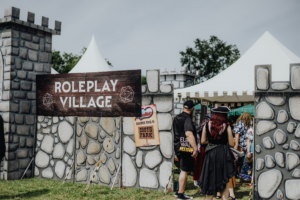 Roleplay Village Besucher erkunden das Roleplay Village auf dem Elbenwald Festival, umgeben von festlichen Ständen und einer mittelalterlichen Kulisse. Die Szene zeigt Teilnehmer in kreativen Kostümen, die die Atmosphäre des Festivals genießen.