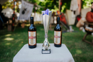 Die Roten Adler vs. 501st Trophäe der Elbenwald Olympiade mit zwei Flaschen Rotwein "Roter Wikinger" auf einem Tisch, umgeben von einer grünen Wiese und dekorativen Elementen im Hintergrund, die die festliche Atmosphäre des Elbenwald Festivals widerspiegeln.