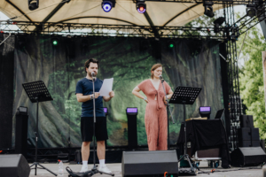 Schatten der Horkruxe by MooEntertainment Zwei Moderatoren stehen auf der Bühne des Elbenwald Festivals, während sie in ein Mikrofon sprechen. Im Hintergrund ist eine grüne Kulisse zu sehen. Die Veranstaltung kombiniert Musik und Nerdkultur und zieht ein kreatives Publikum an.
