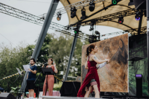 Schatten der Horkruxe by MooEntertainment Künstlerin auf der Bühne des Elbenwald Festivals, die ein großes Gemälde kreiert. Im Hintergrund stehen zwei Moderatoren mit Mikrofonen, während die Menge gespannt zuschaut. Die Bühne ist mit bunten Lichtern und einem großen Leinwandbild dekoriert.