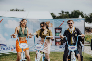 Drei Festivalbesucher am Elbenwald Festival posieren auf Fahrrädern vor einem bunten Hintergrund mit Eiscreme-Werbung. Die Veranstaltung kombiniert Musik, Cosplay und eine lebendige Atmosphäre für Fans von Popkultur und Nerd-Entertainment.