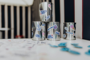 Scoops Ahoy Bunte Eiseimer mit Aufklebern von "Scoops & Ahoy" stehen auf einem Tisch, umgeben von bunten Plättchen. Die dekorativen Eimer sind Teil des Elbenwald Festivals, das Musik und Nerd-Kultur vereint.