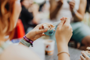 Scoops Ahoy Eine Person hält einen Becher Eis mit einem kleinen Löffel in der Hand, während sie das Elbenwald Festival besucht. Im Hintergrund sind weitere Festivalbesucher zu sehen, die die Atmosphäre genießen. Das Handgelenk ziert ein Festival-Armband.