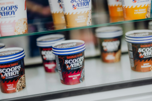 Scoops Ahoy Verschiedene Eissorten von "Scoops Ahoy" aus "Stranger Things" in einem Kühlschrank. Zu sehen sind Geschmacksrichtungen wie Triple Choc Shadow, Raspberry Rift und Spicy Cinnamon. Perfekt für Fans von Nerdkultur und leckeren Snacks beim Elbenwald Festival.