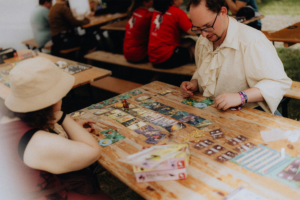 Zwei Teilnehmer spielen ein Brettspiel auf dem Elbenwald Festival, umgeben von weiteren festlichen Aktivitäten und Menschen in nerdiger Kleidung. Auf dem Tisch liegen bunte Spielkarten und Figuren, die das kreative Ambiente des Festivals unterstreichen.