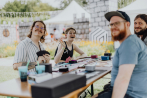 Freunde spielen Karten in einem bunten Outdoor-Bereich beim Elbenwald Festival. Im Hintergrund sind Zelte und eine festliche Atmosphäre sichtbar, die das kreative und gesellige Ambiente des Festivals unterstreichen.