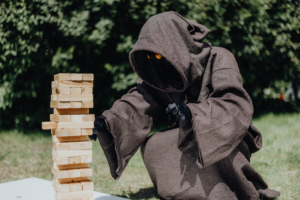 Ein kostümierter Teilnehmer in einem braunen Umhang spielt Jenga auf dem Elbenwald Festival. Die Figur hat leuchtend orange Augen und konzentriert sich darauf, einen Holzblock vorsichtig zu entfernen, während eine grüne Wiese im Hintergrund sichtbar ist.