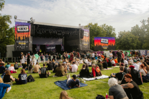 Bühne des Elbenwald Festivals mit Besuchern, die auf dem Gras sitzen. Im Hintergrund sind farbenfrohe Banner und ein großes Display zu sehen, während die Menge auf die Darbietungen wartet. Das Festival vereint Musik und Nerdkultur in einer einzigartigen Atmosphäre.