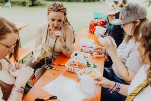 Stitchcraft Teilnehmerinnen beim Elbenwald Festival, die an einem kreativen Stick-Workshop teilnehmen. Auf einem orangefarbenen Tisch sind Stickmaterialien und Werkzeuge verteilt, während die Frauen konzentriert an ihren Projekten arbeiten.