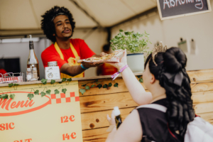 Surfer Boy Pizza Ein Verkäufer reicht einer Festivalbesucherin eine Pizza an einem Essensstand beim Elbenwald Festival. Im Hintergrund sind die Menüpreise sichtbar, umgeben von einer rustikalen Holztheke und grünen Pflanzen. Die Atmosphäre ist lebhaft und einladend.