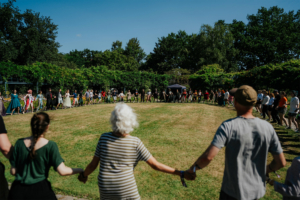 Historische Tänze Menschen in bunten Kostümen tanzen Hand in Hand auf dem Elbenwald Festival, umgeben von grünen Bäumen und strahlend blauem Himmel. Die fröhliche Atmosphäre spiegelt die Vielfalt der Teilnehmer und die Gemeinschaft der Nerd-Kultur wider.