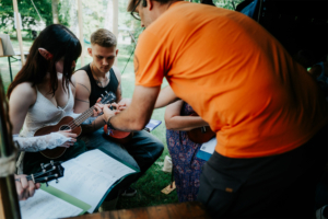 Teilnehmer des Elbenwald Festivals lernen gemeinsam Ukulele spielen. Eine Person mit Elfenohren spielt, während andere Anleitung erhalten. Notenblätter liegen auf dem Tisch, um die musikalische Interaktion zu unterstützen.