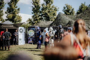 Olympiade Die Roten Adler vs. 501st Legion Besucher des Elbenwald Festivals in Kostümen stehen vor verschiedenen Ständen, darunter eine Kommandozentrale. Im Hintergrund sind mehrere Charaktere aus der Science-Fiction-Welt zu sehen, die die lebendige Atmosphäre des Festivals unterstreichen.