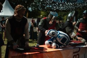 Olympiade Die Roten Adler vs. 501st Legion Zwei verkleidete Teilnehmer am Elbenwald Festival präsentieren ein gehäkeltes R2-D2-Spielzeug auf einem Tisch, während im Hintergrund weitere Festivalbesucher in Kostümen zu sehen sind. Die Szenerie vereint Fantasie und Gemeinschaft.