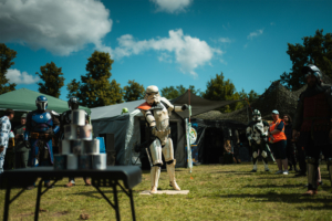 Olympiade Die Roten Adler vs. 501st Legion Cosplay-Teilnehmer in verschiedenen Kostümen, darunter Stormtrooper und Mandalorianer, stehen auf dem Elbenwald Festival. Im Vordergrund sind Stapel von Dosen auf einem Tisch zu sehen, während die Festivalbesucher die Atmosphäre genießen.