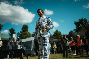 Olympiade Die Roten Adler vs. 501st Legion Ein Mann in einem auffälligen Anzug mit R2-D2-Muster hält ein Spielzeug-Hühnchen in der Hand. Im Hintergrund sind Menschen in Kostümen und Festivalzelte zu sehen, was die lebhafte Atmosphäre des Elbenwald Festivals unterstreicht.