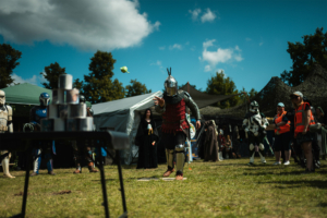 Olympiade Die Roten Adler vs. 501st Legion Ein kostümierter Teilnehmer wirft während des Elbenwald Festivals eine grüne Kugel auf einen Stapel Dosen. Im Hintergrund sind weitere Cosplayer und festivaltypische Zelte zu sehen, die die lebendige Atmosphäre des Events unterstreichen.