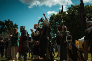 Olympiade Die Roten Adler vs. 501st Legion Menschen in Kostümen feiern enthusiastisch auf dem Elbenwald Festival, umgeben von einer fröhlichen Atmosphäre. Ein Ritter mit Helm und andere Teilnehmer zeigen ihre Begeisterung während einer Veranstaltung im Freien, unter blauem Himmel und Sonnenschein.
