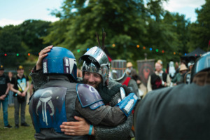 Olympiade Die Roten Adler vs. 501st Legion Zwei verkleidete Teilnehmer des Elbenwald Festivals umarmen sich herzlich, während im Hintergrund andere Kostümierte und festliche Lichter zu sehen sind. Die Szene strahlt Gemeinschaft und Freude in einer nerdigen Festivalatmosphäre aus.