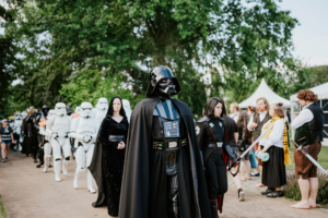 501st Legion Cosplay-Parade beim Elbenwald Festival: Darth Vader führt eine Gruppe von Stormtroopers und weiteren Charakteren durch den Festivalgelände, umgeben von Bäumen und Zelten. Die Teilnehmer verkörpern verschiedene Helden und Schurken aus der Popkultur.