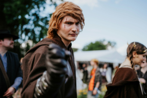 501st Legion Ein junger Mann in einem braunen Umhang mit einer entschlossenen Miene und einem erhobenen Arm, umringt von anderen kostümierten Teilnehmern des Elbenwald Festivals, das Musik und Nerd-Kultur vereint, unter blauem Himmel und Bäumen.