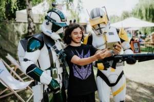 501st Legion Ein junger Mann macht ein Selfie mit zwei Kostümierten in Rüstungen auf dem Elbenwald Festival. Die Szene zeigt die kreative Verbindung von Musik und Nerdkultur, umgeben von Festivalatmosphäre und begeisterten Besuchern.